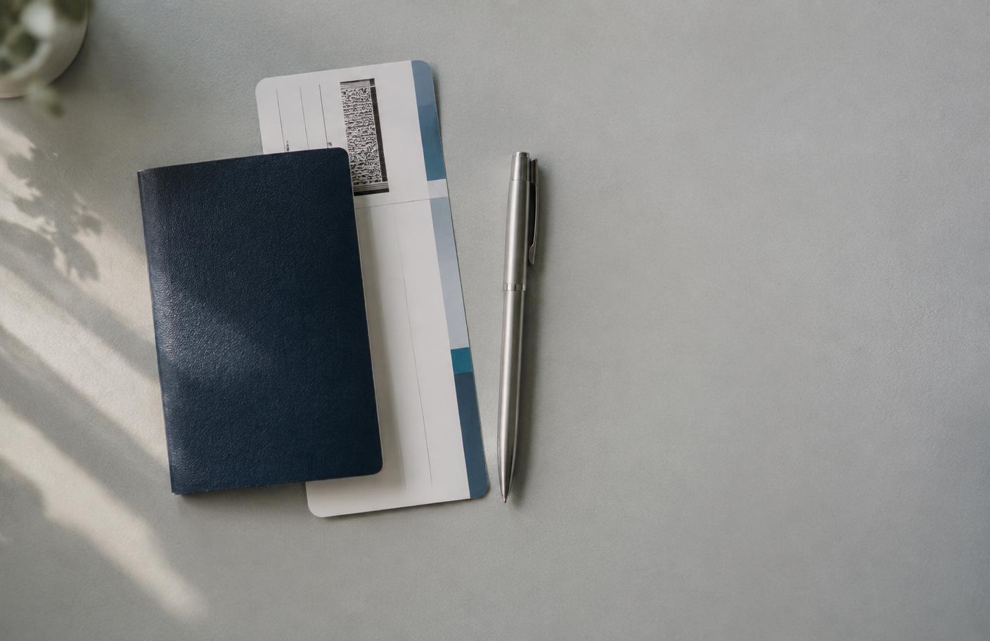 A boarding pass, a passport and a pen laid out on a plain desk in natural window light.